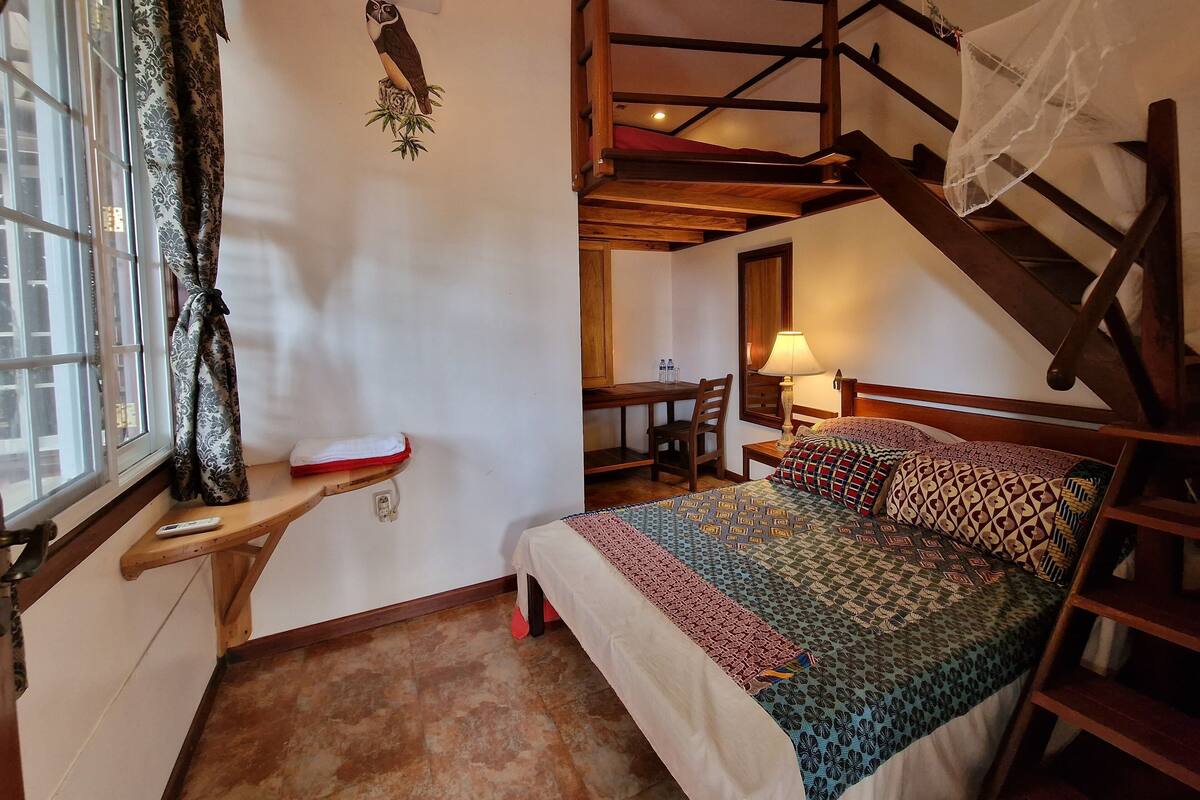 A bedroom with a bed, a desk, a chair, a window with curtains, a staircase, and an air conditioning unit.