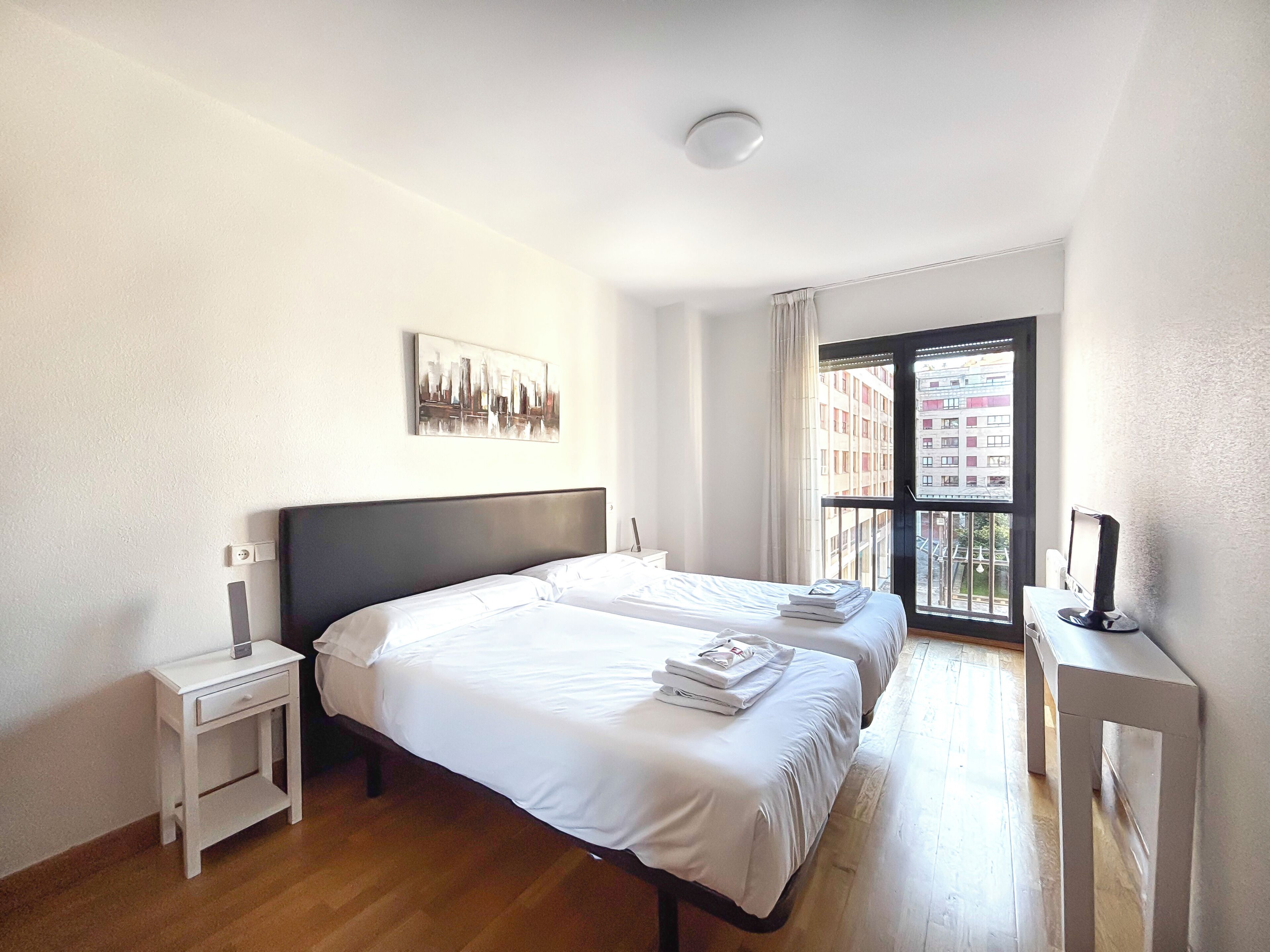 Habitación de hotel con dos camas, una mesita de noche, un escritorio con computadora, una ventana con vistas a edificios y una lámpara en el techo.