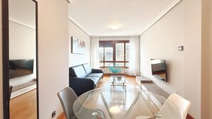 A modern living room with a glass table, a black sofa, a TV, and a window with curtains. - Apartamentos Gestión de Alojamientos (Pamplona)