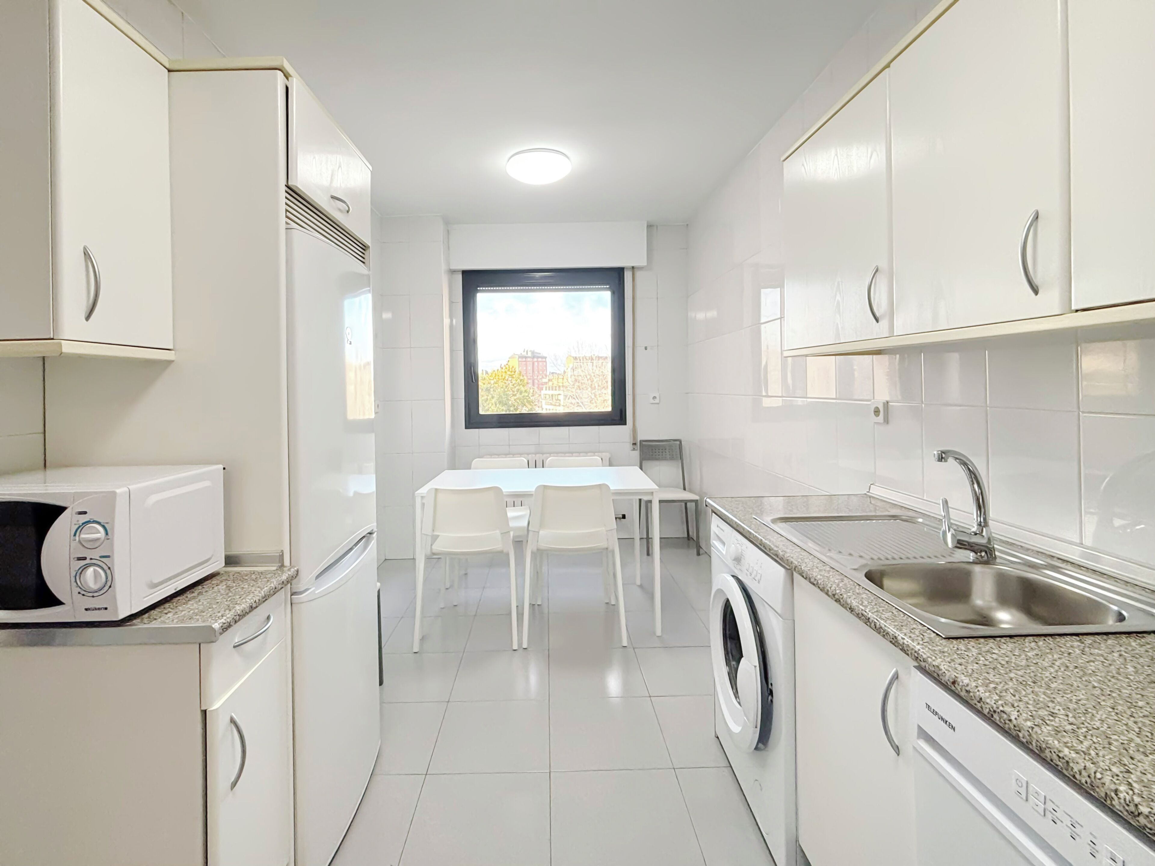 A compact kitchen with white cabinets, a microwave, a washing machine, and a dining area with a table and chairs.