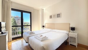 A hotel room with two beds, a balcony with a city view, a desk with a computer, and a bedside table with a lamp. - Apartamentos Gestión de Alojamientos (Pamplona)