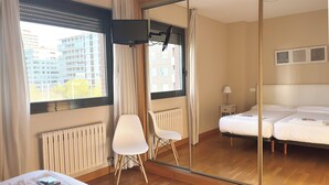 A hotel room with a bed, a chair, a desk, a mirror, and a view of buildings outside. - Apartamentos Gestión de Alojamientos (Pamplona)