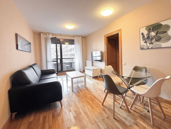 A modern living room with a black leather sofa, a glass dining table with chairs, a white TV on the wall, and a balcony with a view of buildings.