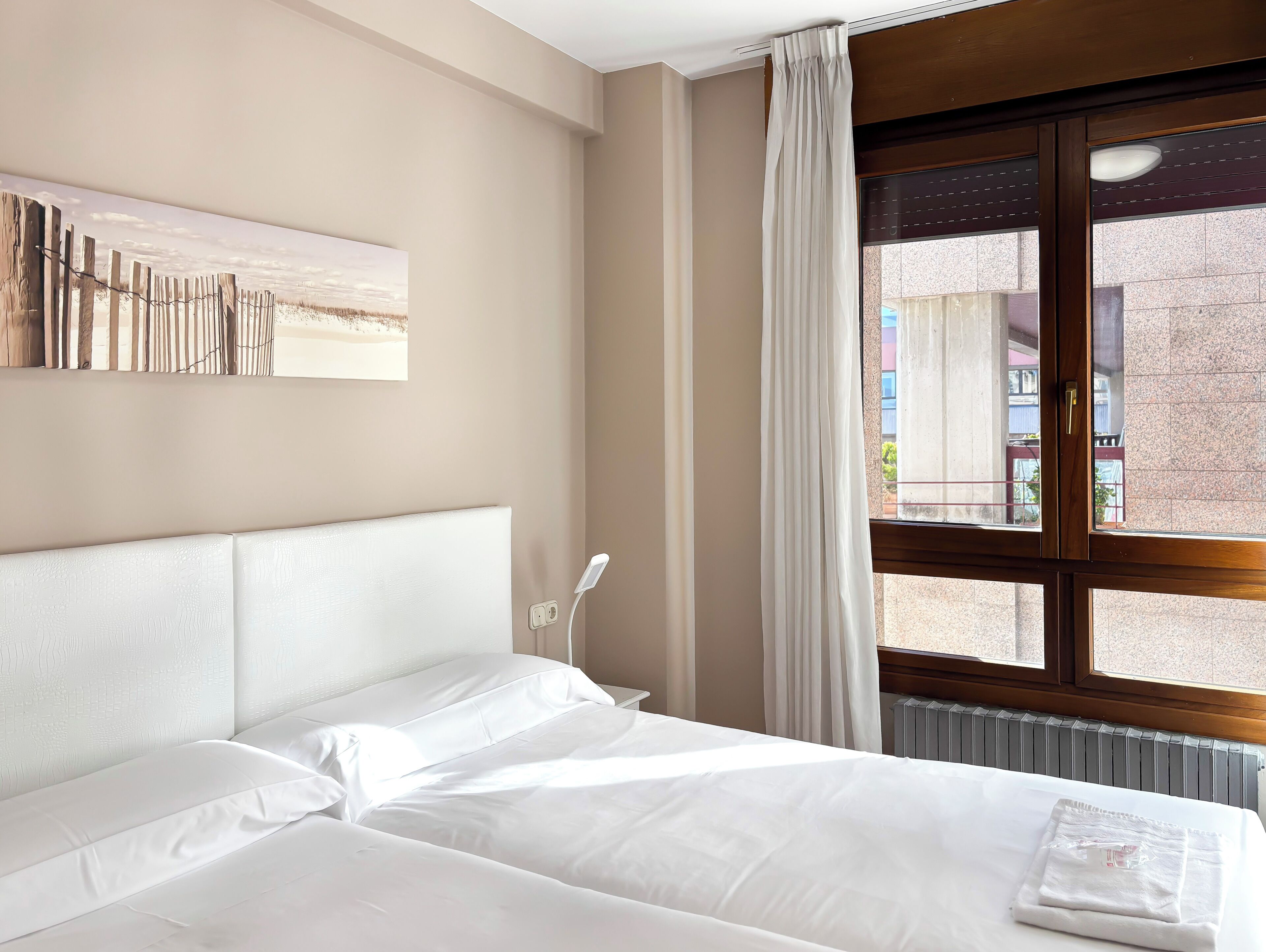A hotel room with a bed, a bedside table, a window with curtains, a wall-mounted artwork, and a radiator.