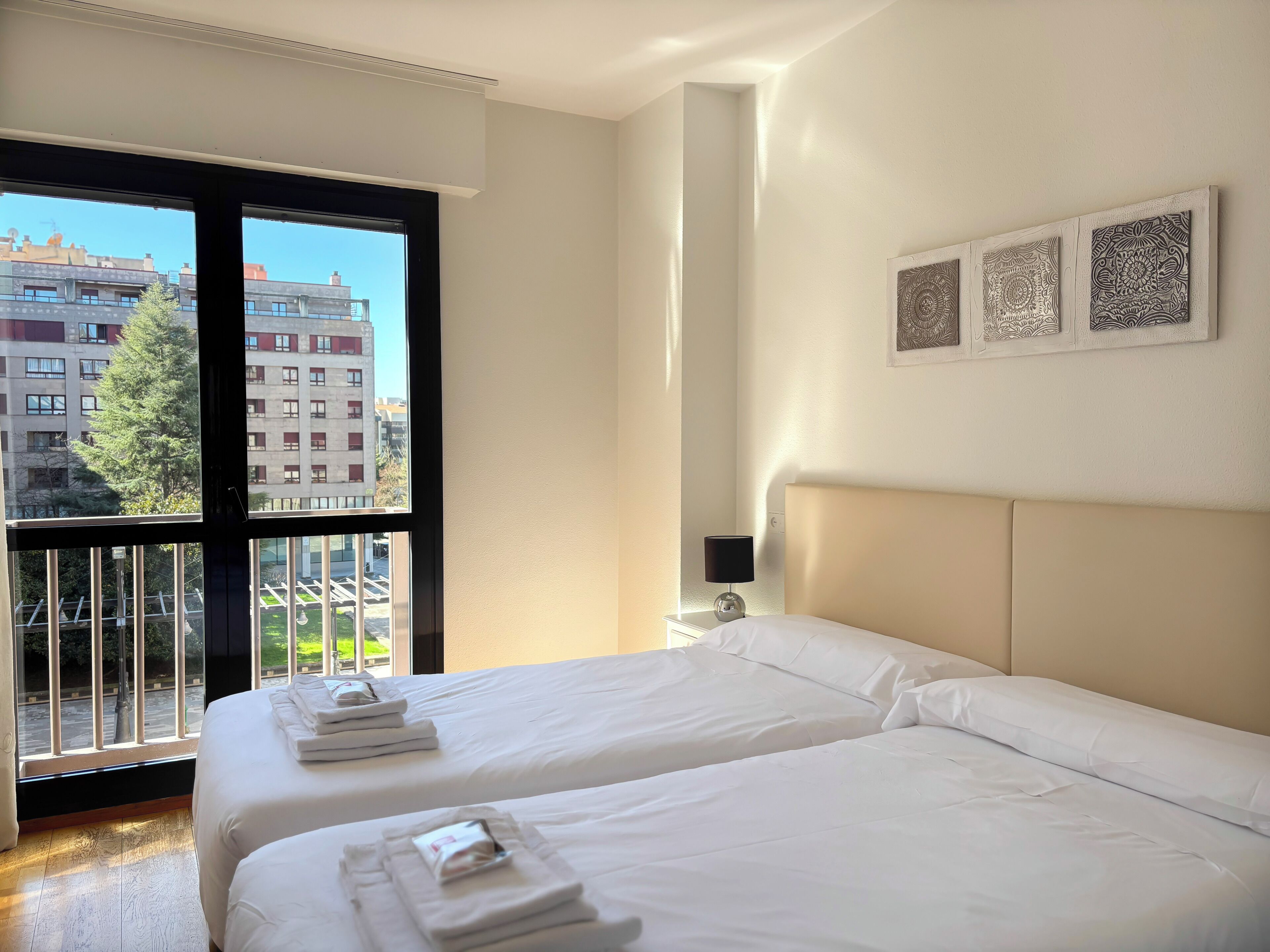 Habitació d'hotel amb dos llits, una finestra gran que mostra edificis, una taula de nit amb una làmpada i una obra d'art penjada a la paret.