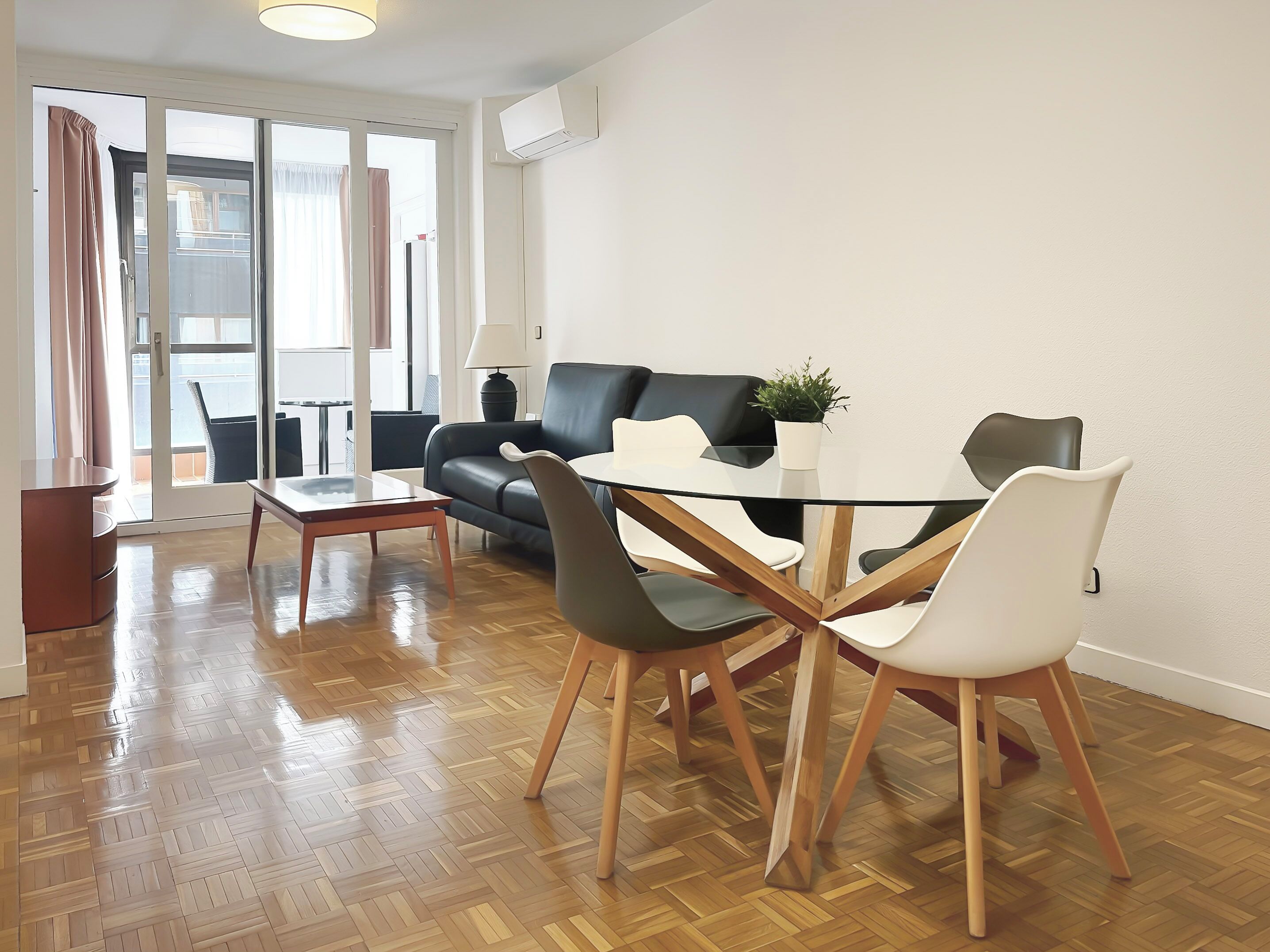 A modern living room with a glass dining table, black and white chairs, a leather sofa, and a potted plant.