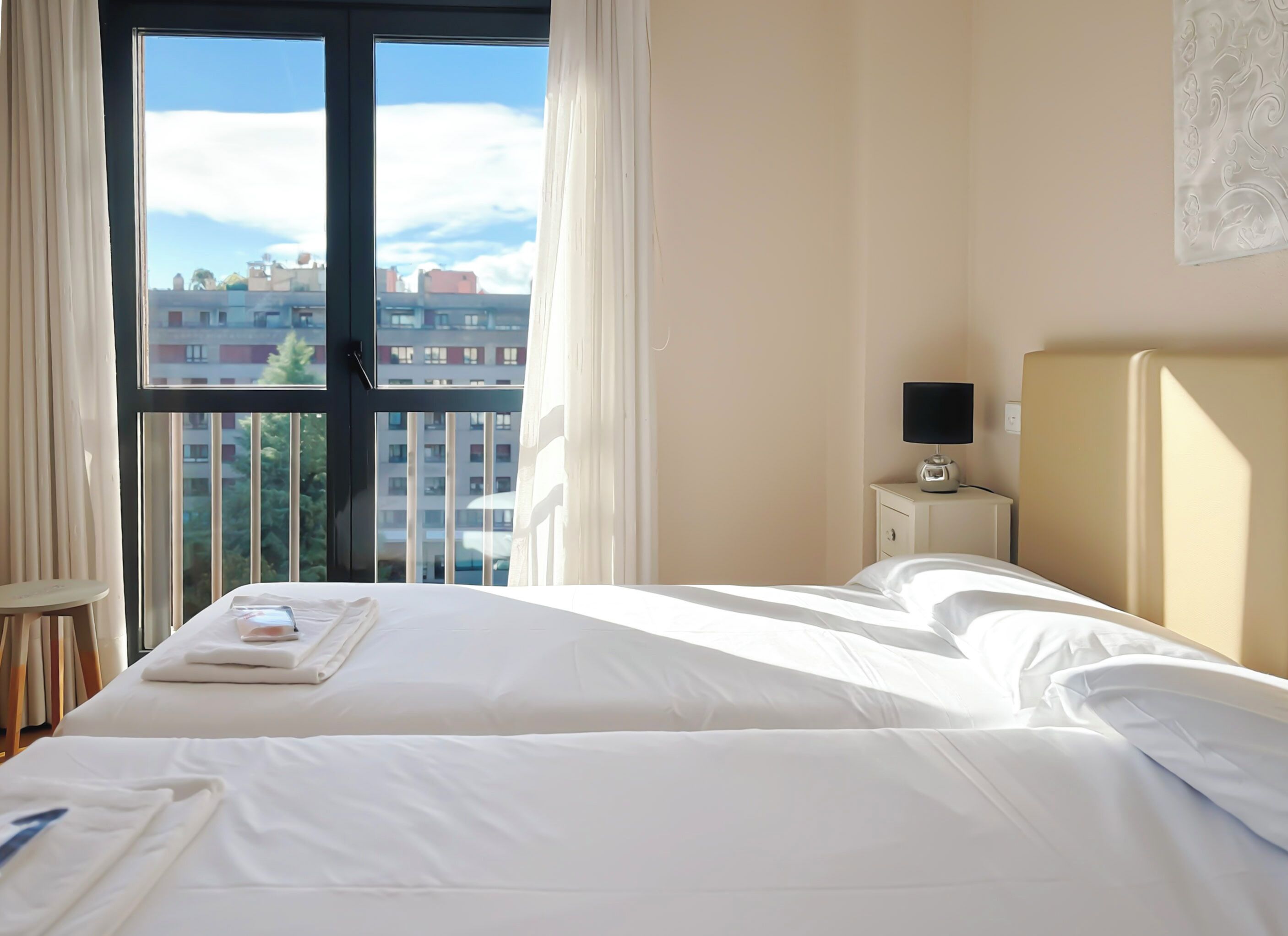 Habitación de hotel con un ventanal grande, una cama, una mesita de noche con lámpara y vistas a edificios.