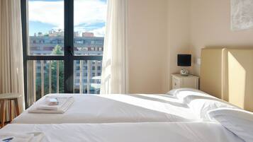 Habitación de hotel con un ventanal grande, una cama, una mesita de noche con lámpara y vistas a edificios.