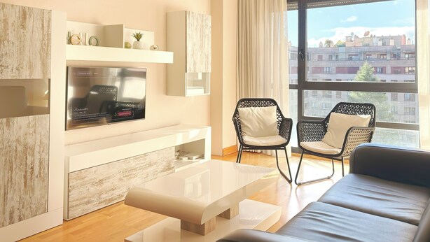 A modern living room with a flat-screen TV, two black wicker chairs, a white sofa, and a large window with a city view. - Apartamentos Gestión de Alojamientos (Pamplona)