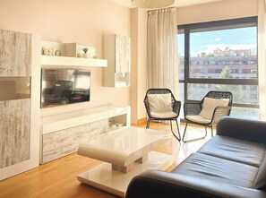 A modern living room with a flat-screen TV, two black wicker chairs, a white sofa, and a large window with a city view. - Apartamentos Gestión de Alojamientos (Pamplona)