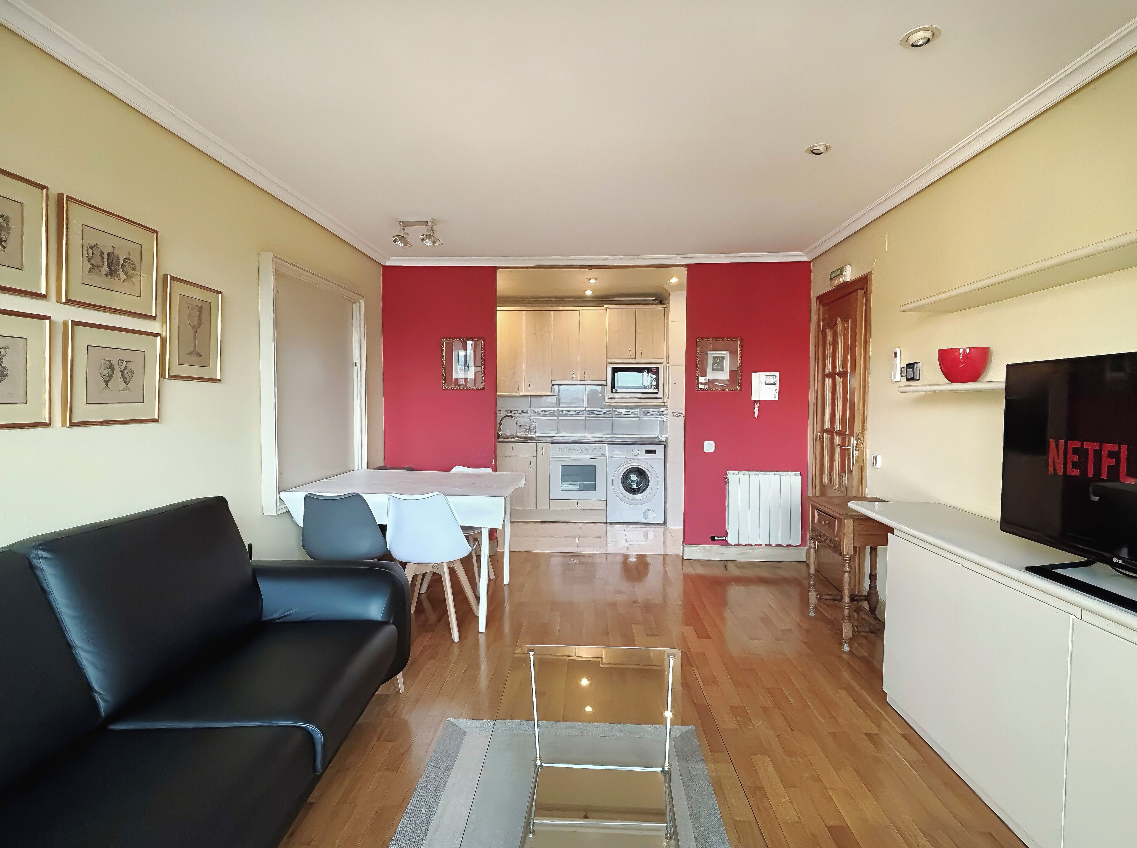 A living room with a black sofa, a dining table with chairs, a kitchen with a microwave, a washing machine, and a TV displaying 'NETFLIX'.