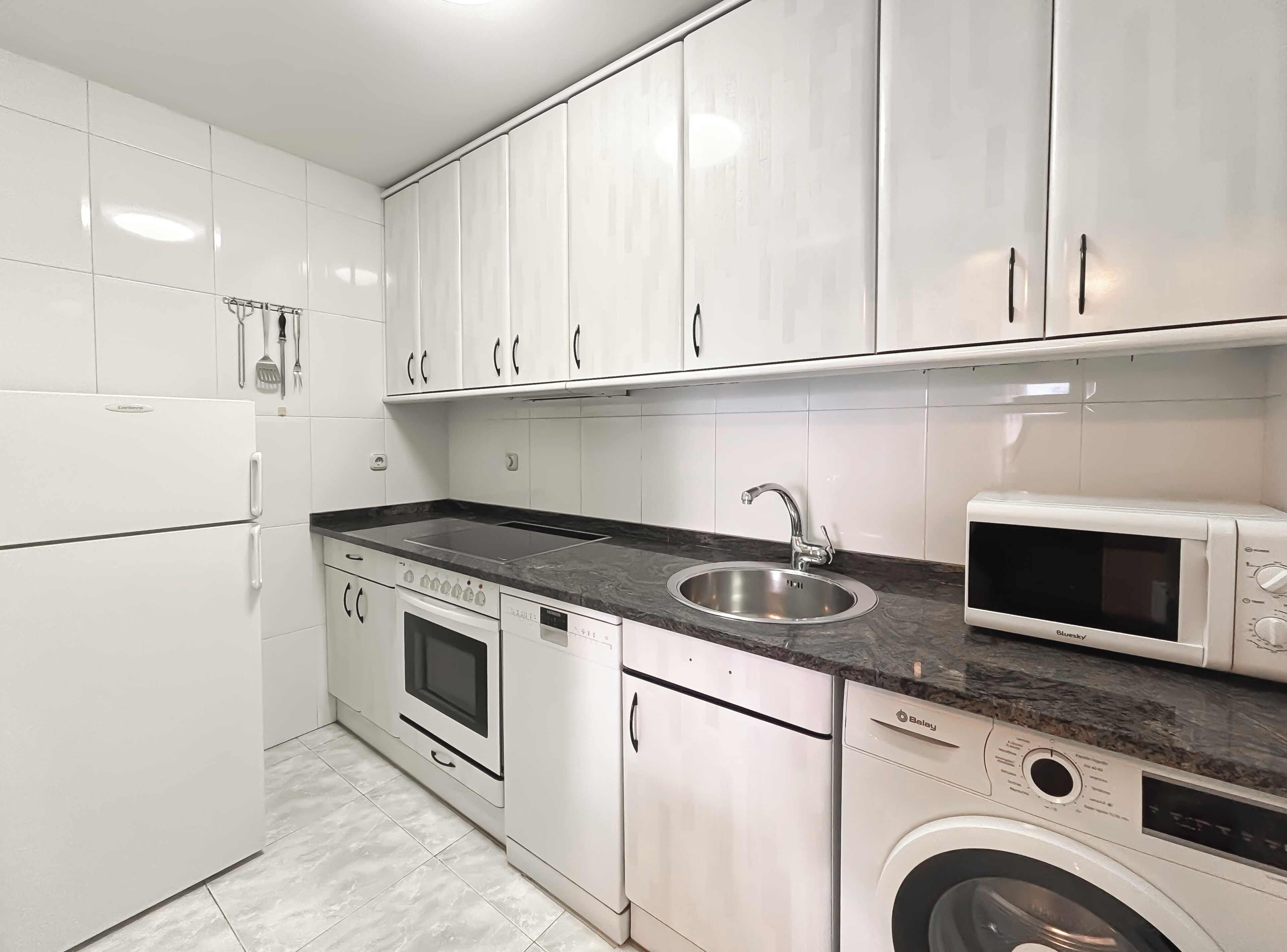 A modern kitchen with white cabinets, a stainless steel sink, a built-in oven, a dishwasher, a microwave, and a washing machine.