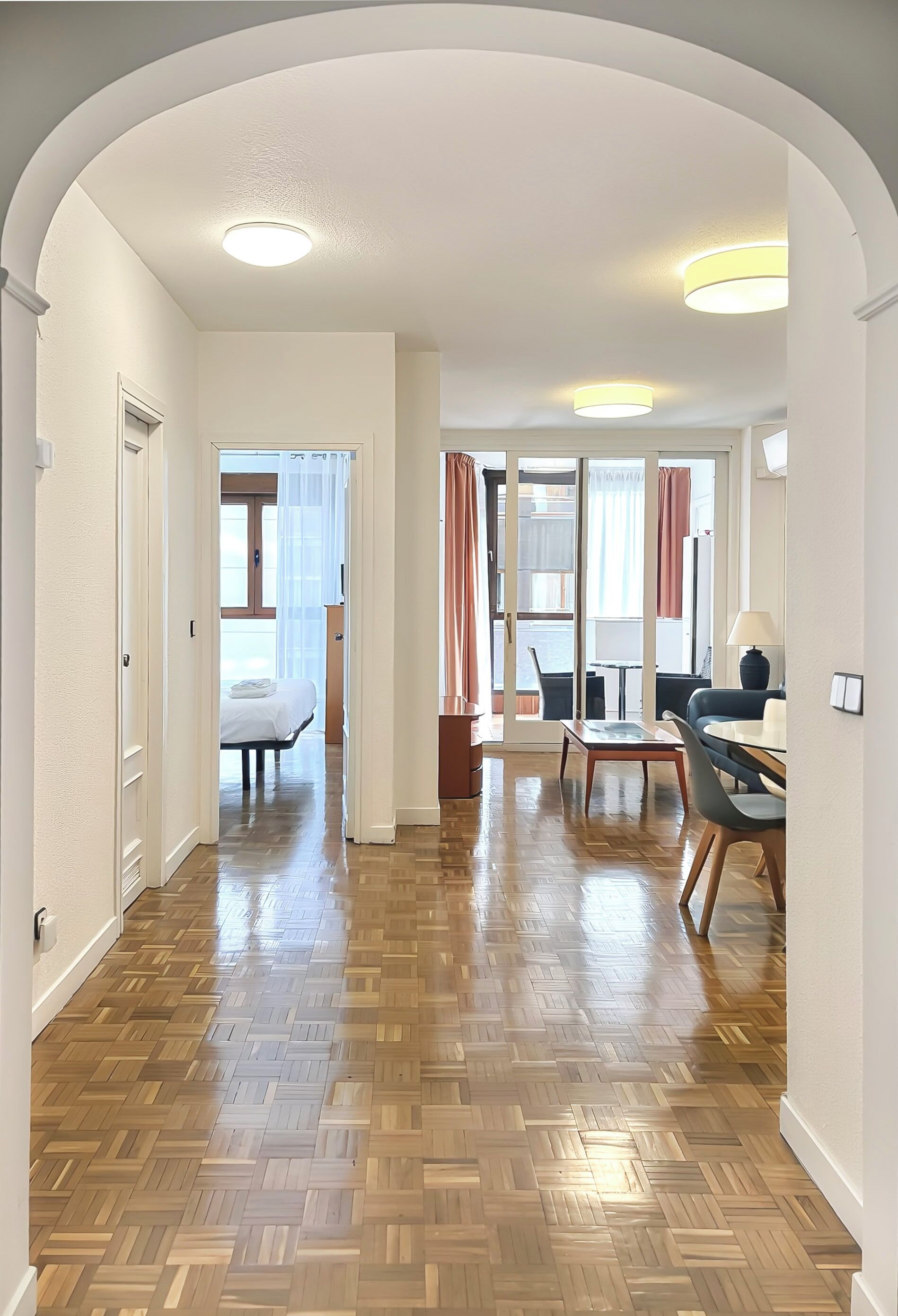 A hallway with wooden flooring, a bed, a dining area with a table and chairs, and a view through a glass door.