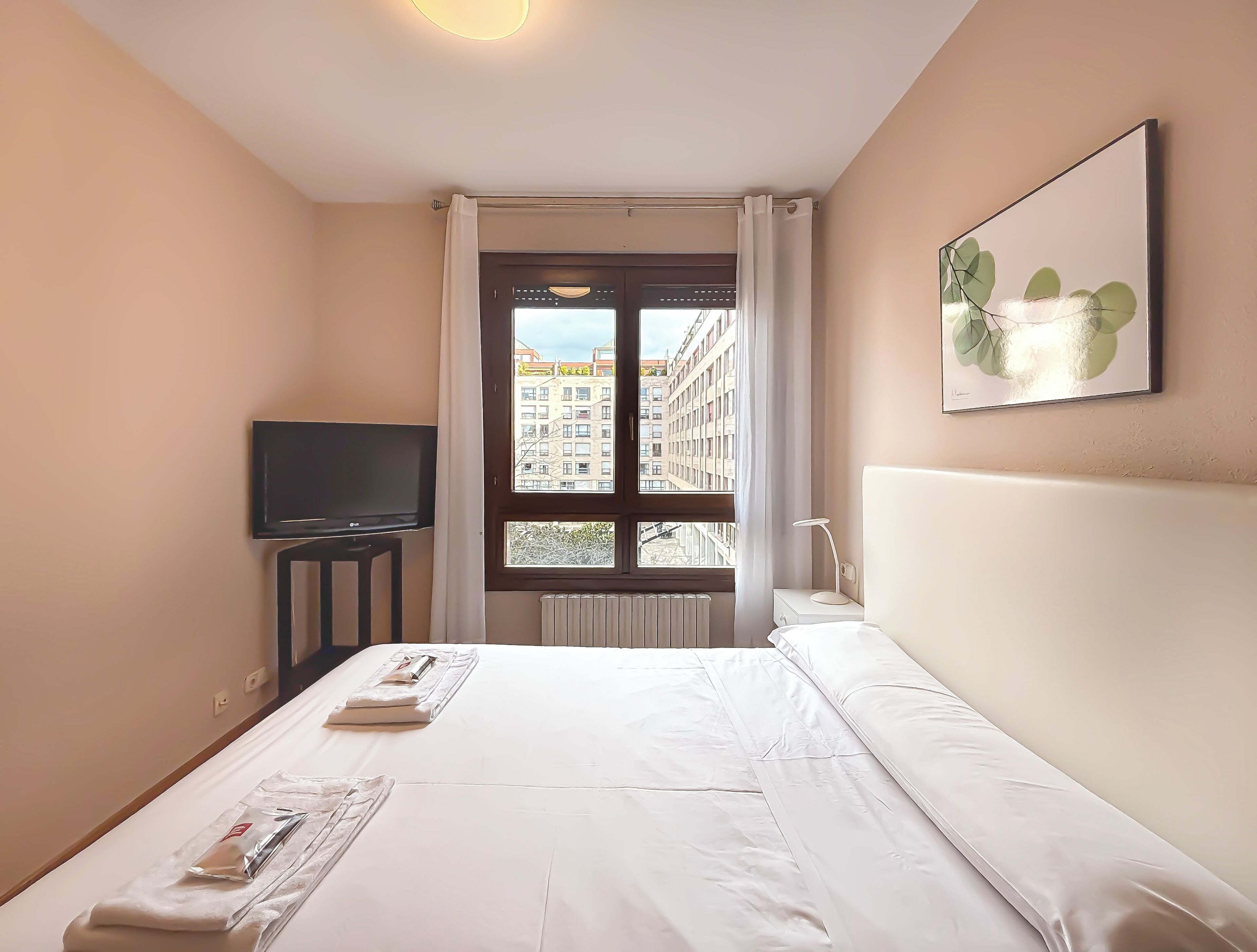 A hotel room with a bed, a television, a window with curtains, and a framed picture on the wall.