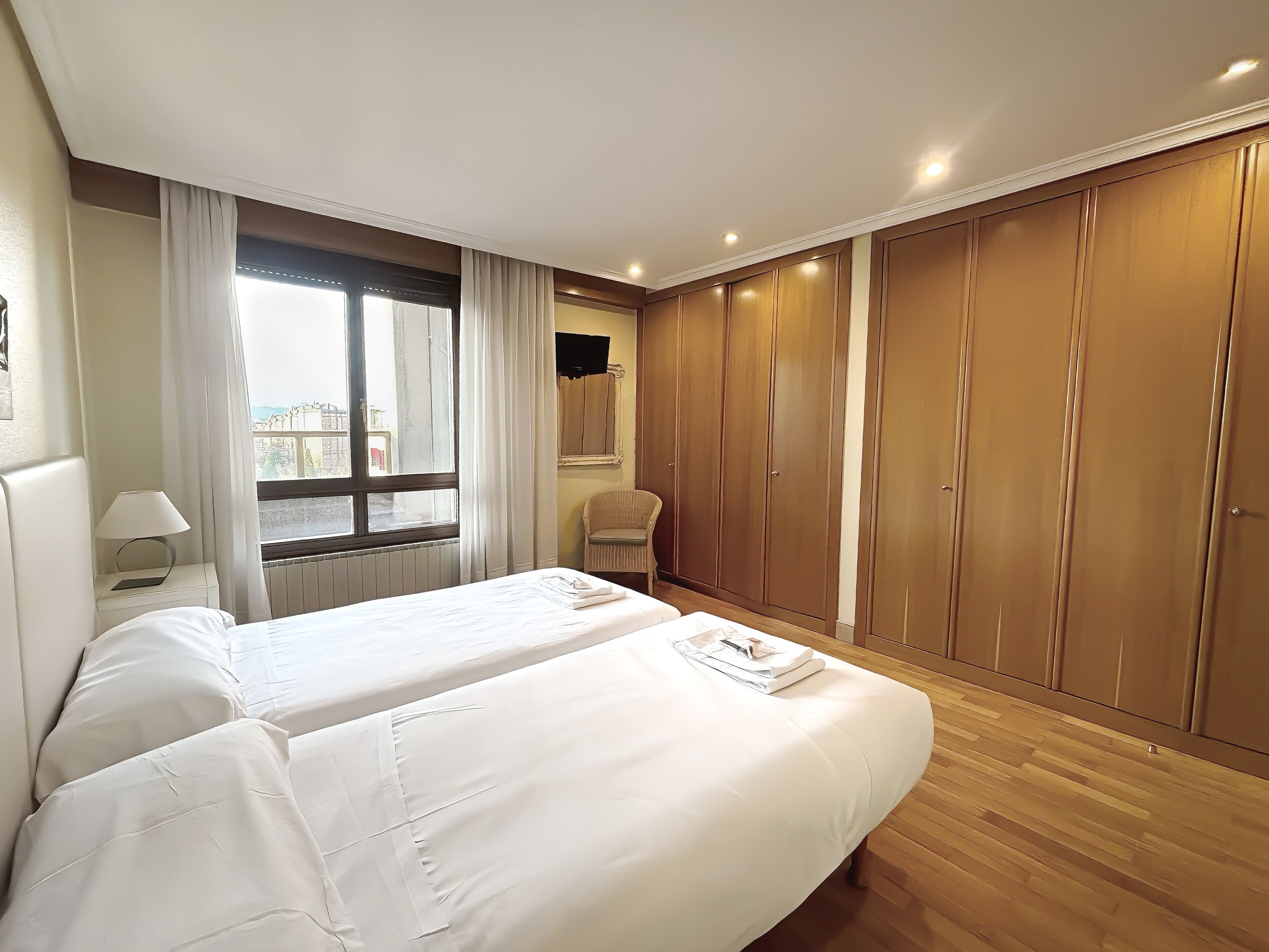 A hotel room with two beds, a window with curtains, a chair, a wardrobe, and a TV mounted on the wall.