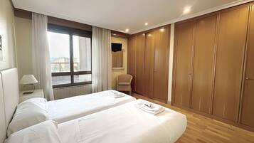 A hotel room with two beds, a window with curtains, a chair, a wardrobe, and a TV mounted on the wall.