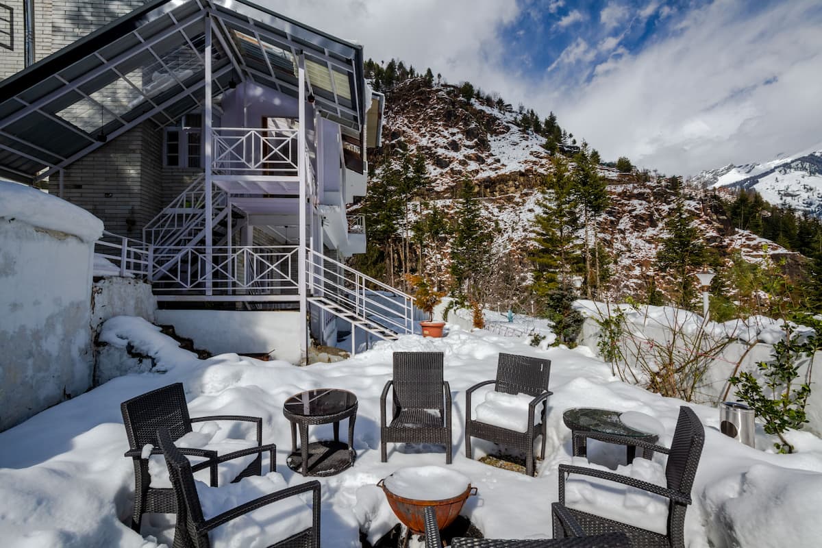 Un espacio al aire libre cubierto de nieve con sillones y mesas de mimbre, una escalera metálica y montañas al fondo.