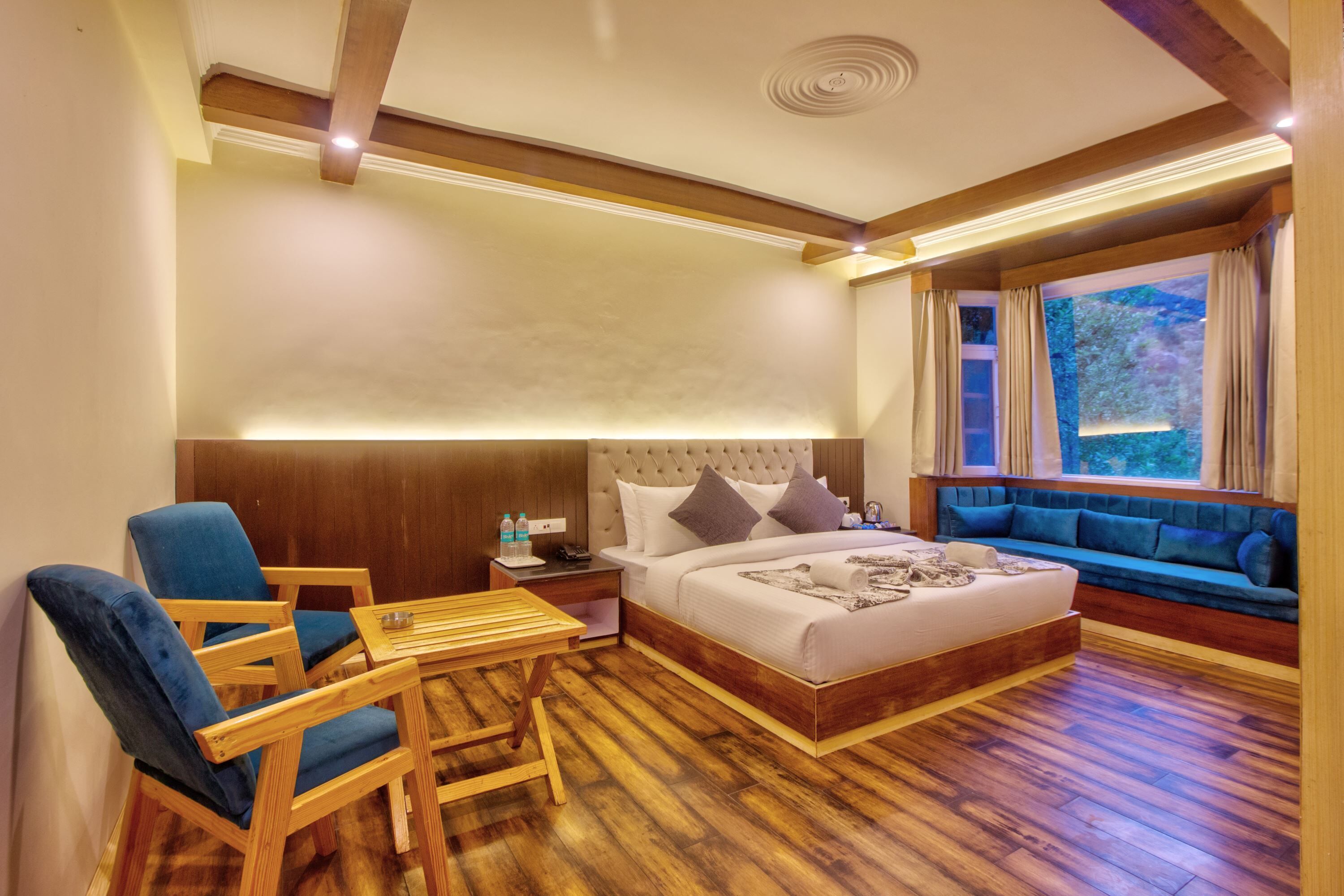 A hotel room with a large bed, a wooden table, blue chairs, a sofa, and a window with curtains.