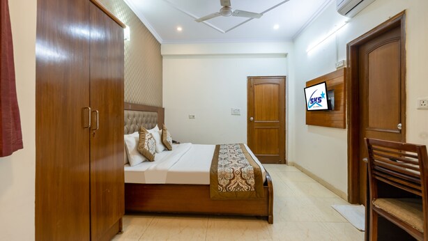 A bedroom with a bed, wardrobe, ceiling fan, TV, and a wooden chair. -  SKS Excel Inn (Gurugram)