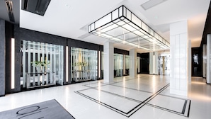 Un moderno vestíbulo de hotel con un diseño limpio y minimalista, que presenta un amplio mostrador de recepción, plantas en macetas y un elemento de iluminación geométrico.