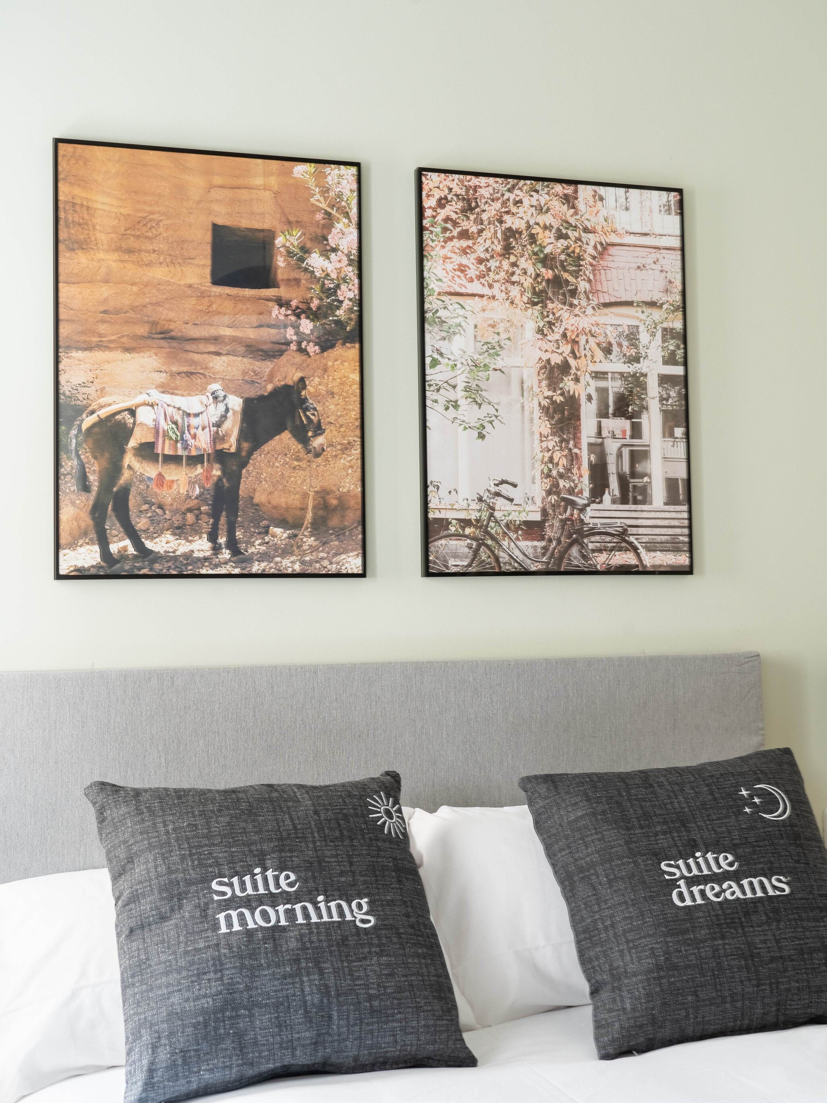 Deux tableaux encadrés sont accrochés au mur, au-dessus d’un lit agrémenté d’oreillers sur lesquels on peut lire les inscriptions « bonne matinée en suite » et « douces nuits en suite ».