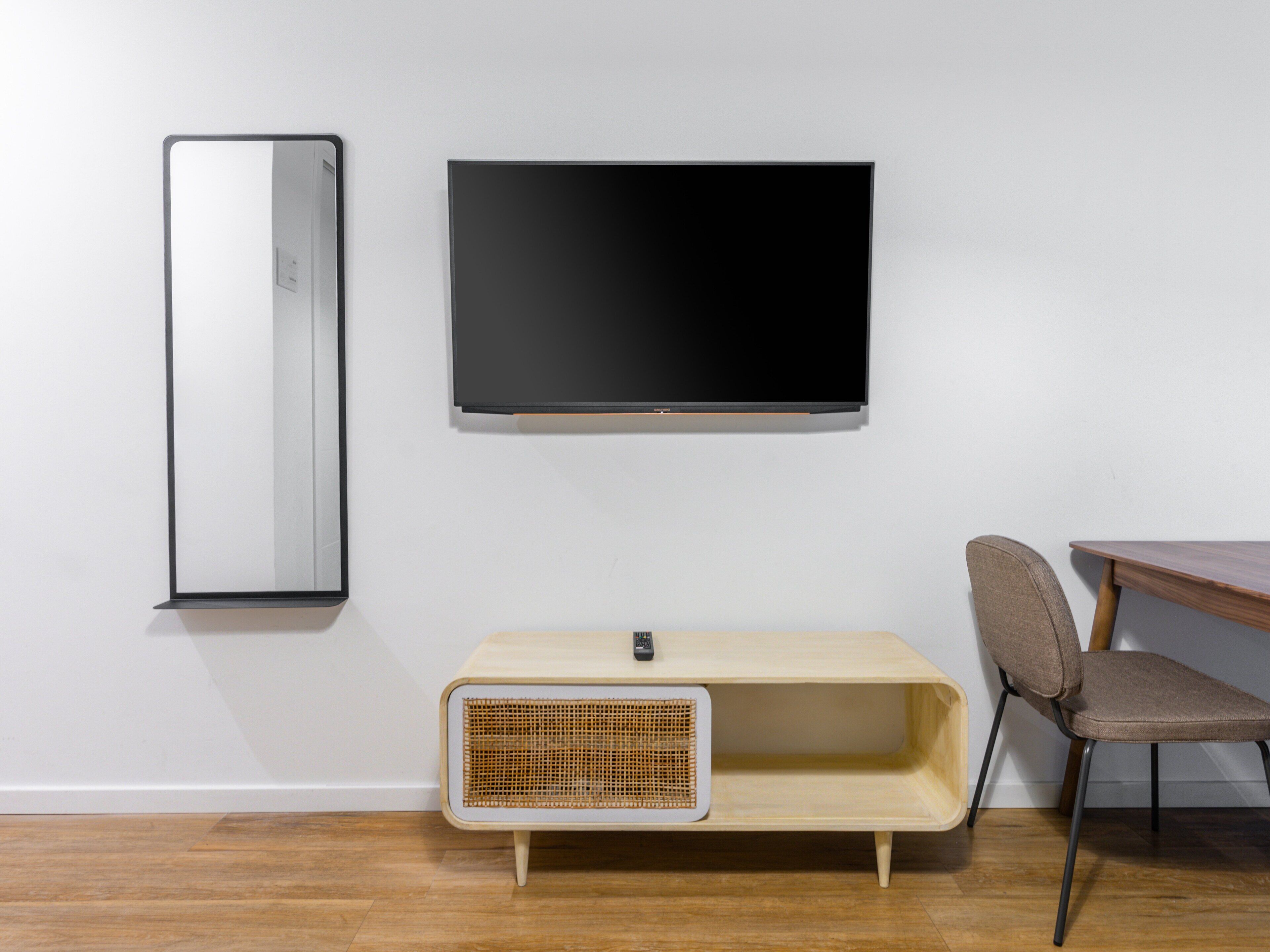 A room with a wall-mounted TV, a mirror, a wooden side table with a wicker basket, and a chair.