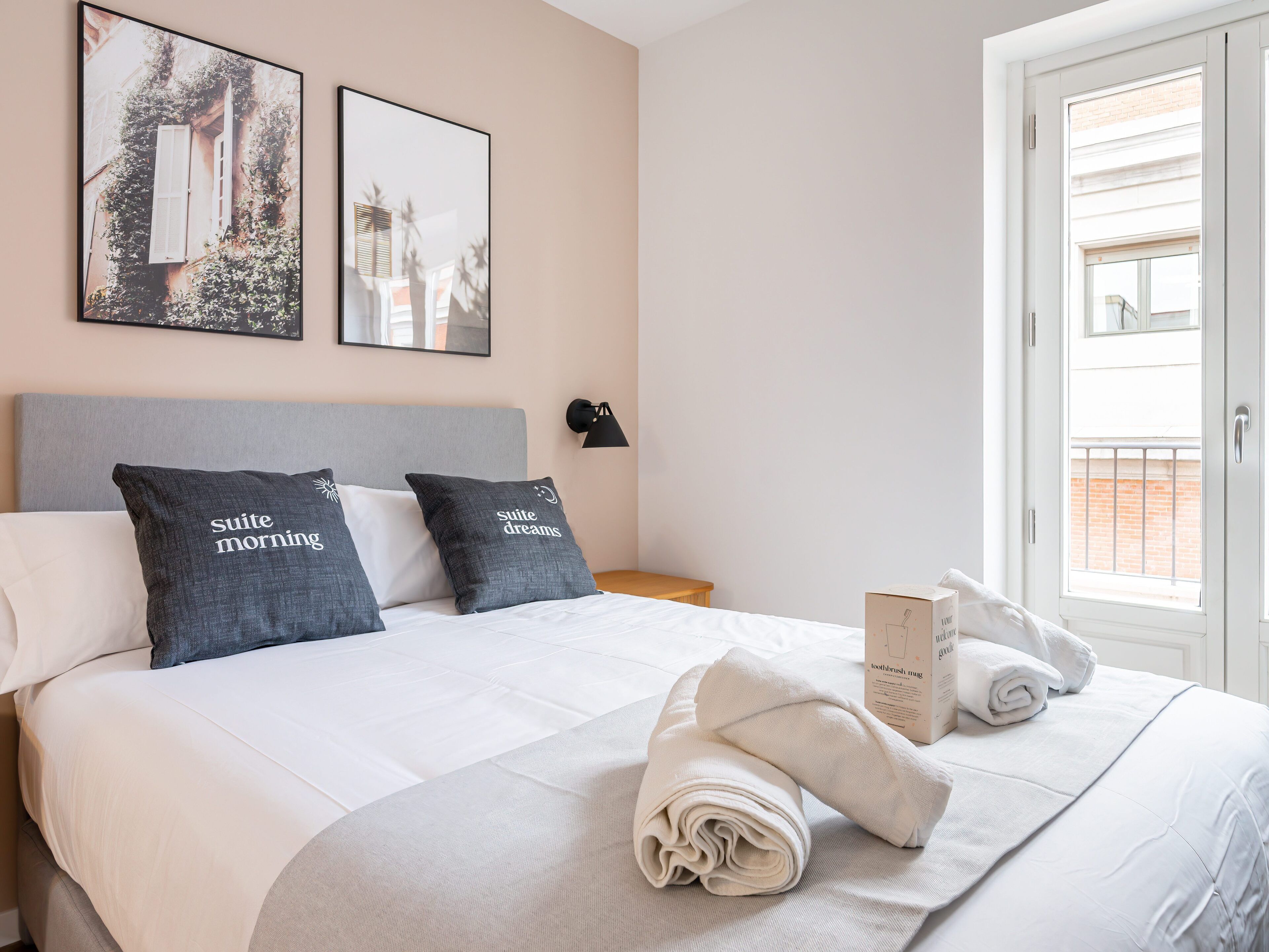 Ein ordentlich bezogenes Bett mit weißer Bettwäsche und zwei Kissen mit Aufschrift, ein Nachttisch, eine Lampe und ein Fenster mit Blick auf Gebäude.