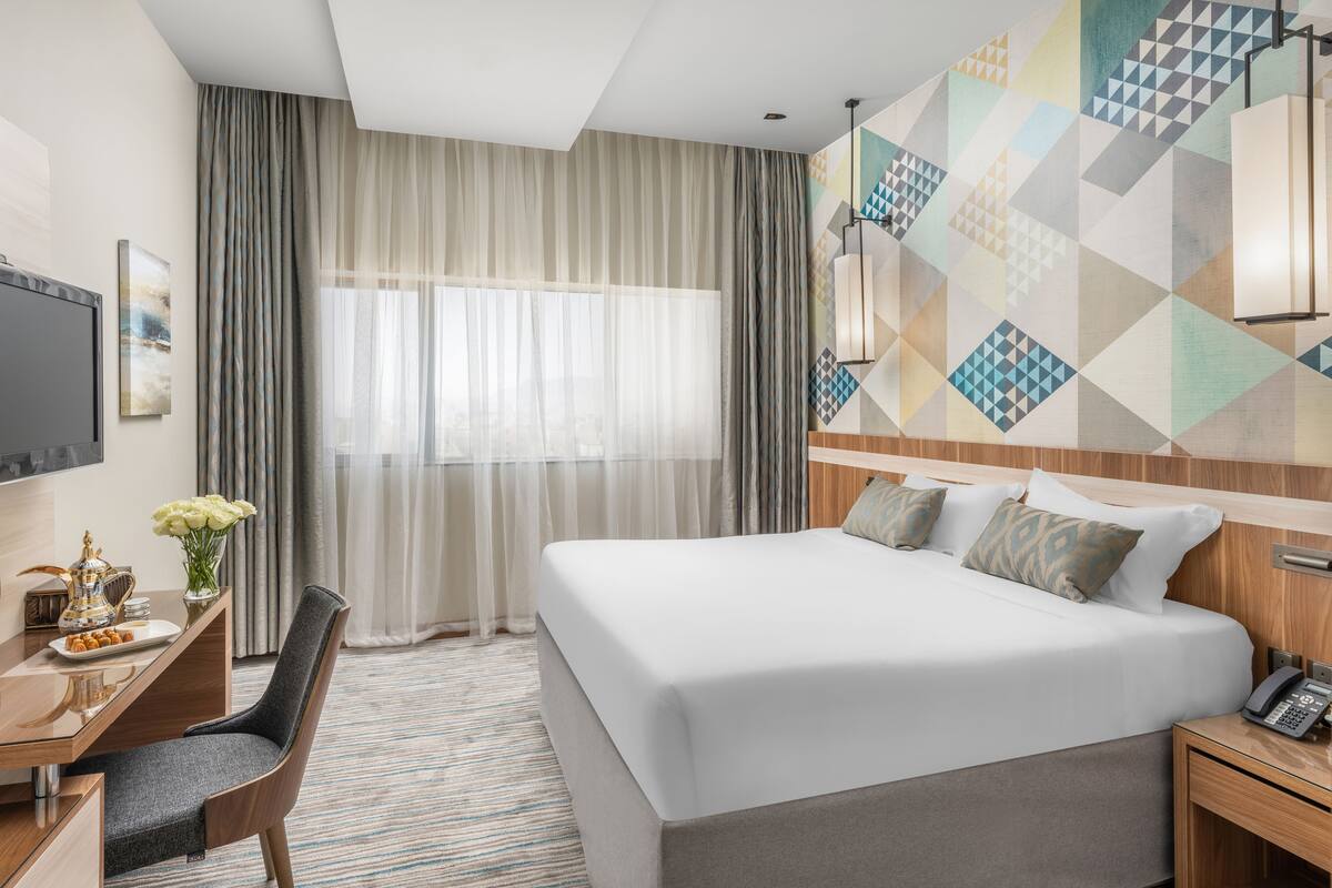 A hotel room with a large bed, a desk with a chair, a TV, and a wall with geometric patterns.