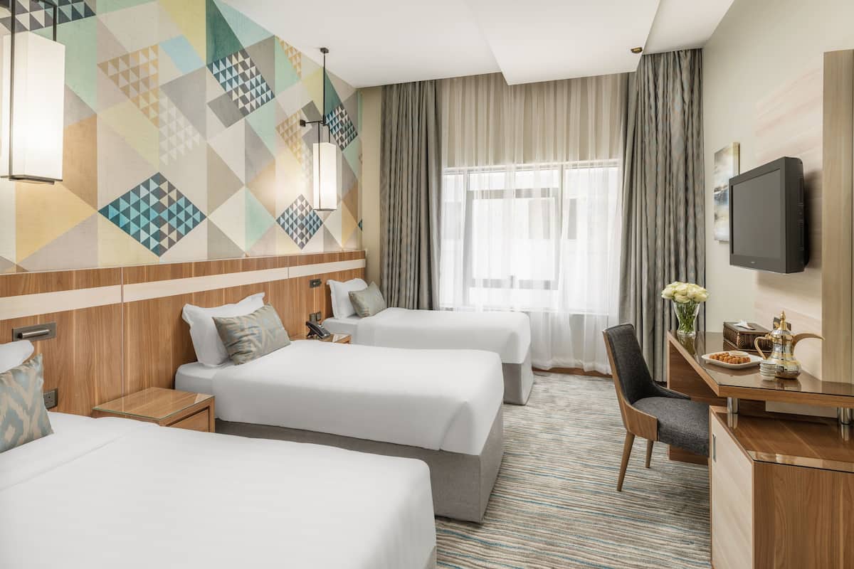 A hotel room with two beds, a desk, a chair, a TV, and a patterned wall.