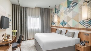A hotel room with a large bed, a desk with a chair, a TV, and a wall with geometric patterns. - Roots Hotel (Makkah)