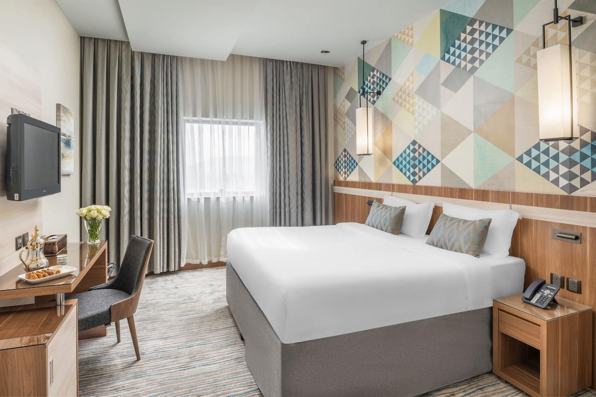 A hotel room with a large bed, a desk with a chair, a TV, and a wall with geometric patterns.