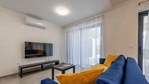 Ein modernes Wohnzimmer mit einem blauen Sofa, einem kleinen Couchtisch, einem flachbildfernseher an der Wand und einem großen Fenster mit durchsichtigen Vorhängen.