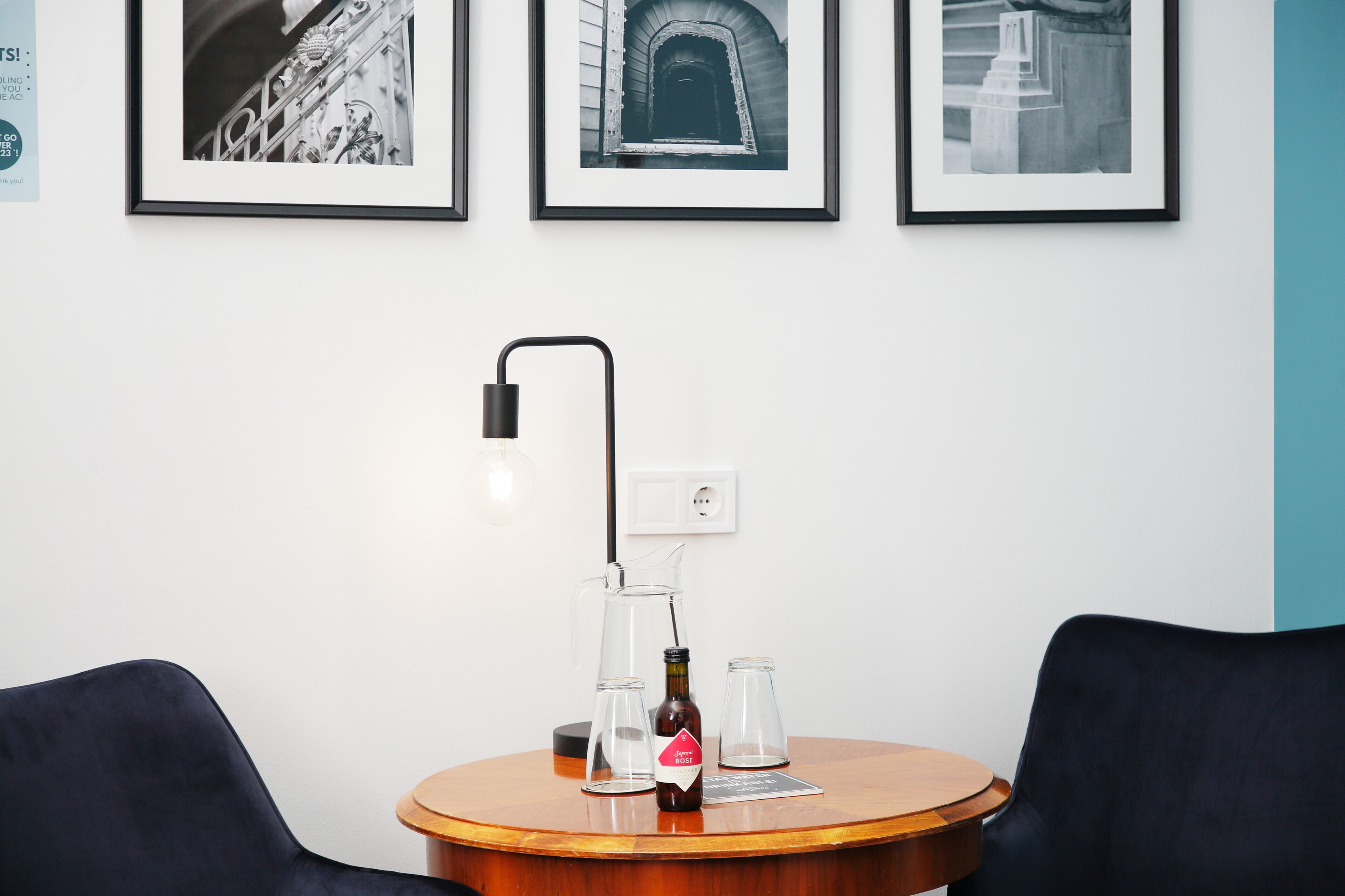 Una mesa redonda de madera con una botella de cerveza, dos vasos vacíos y una lámpara de pie negra. Encima de la mesa, hay tres fotografías en blanco y negro colgadas en la pared.