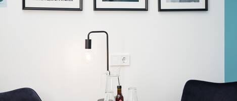 Una mesa redonda de madera con una botella de cerveza, dos vasos vacíos y una lámpara de pie negra. Encima de la mesa, hay tres fotografías en blanco y negro colgadas en la pared.