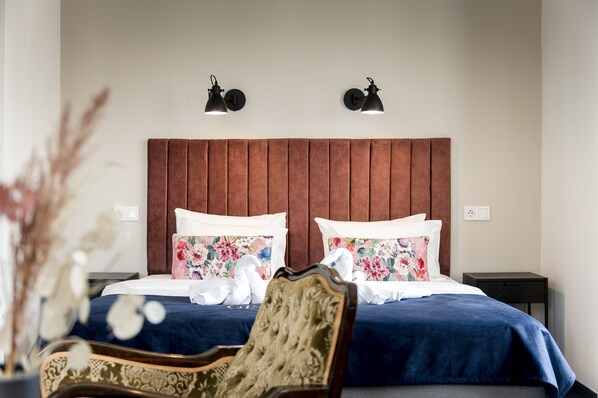 Habitación de hotel con cabecera de madera, dos lámparas de noche, almohadas estampadas, colcha azul y una silla decorativa.