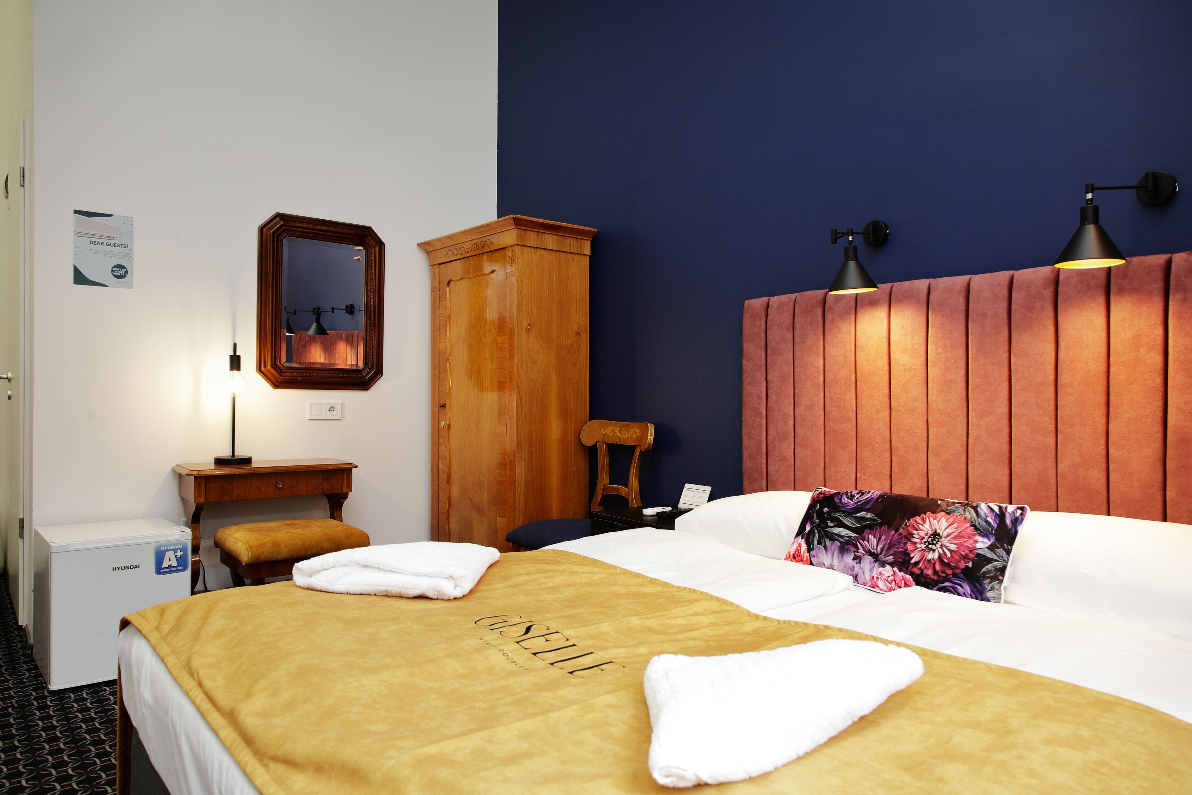 Una camera d'hotel con un letto, un armadio in legno, un comodino, una scrivania, una sedia, uno specchio e una lampada a muro.