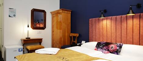 Una camera d'hotel con un letto, un armadio in legno, un comodino, una scrivania, una sedia, uno specchio e una lampada a muro.