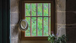 Quarto com paredes de pedra, uma janela, um chapéu e um vaso com flores.
