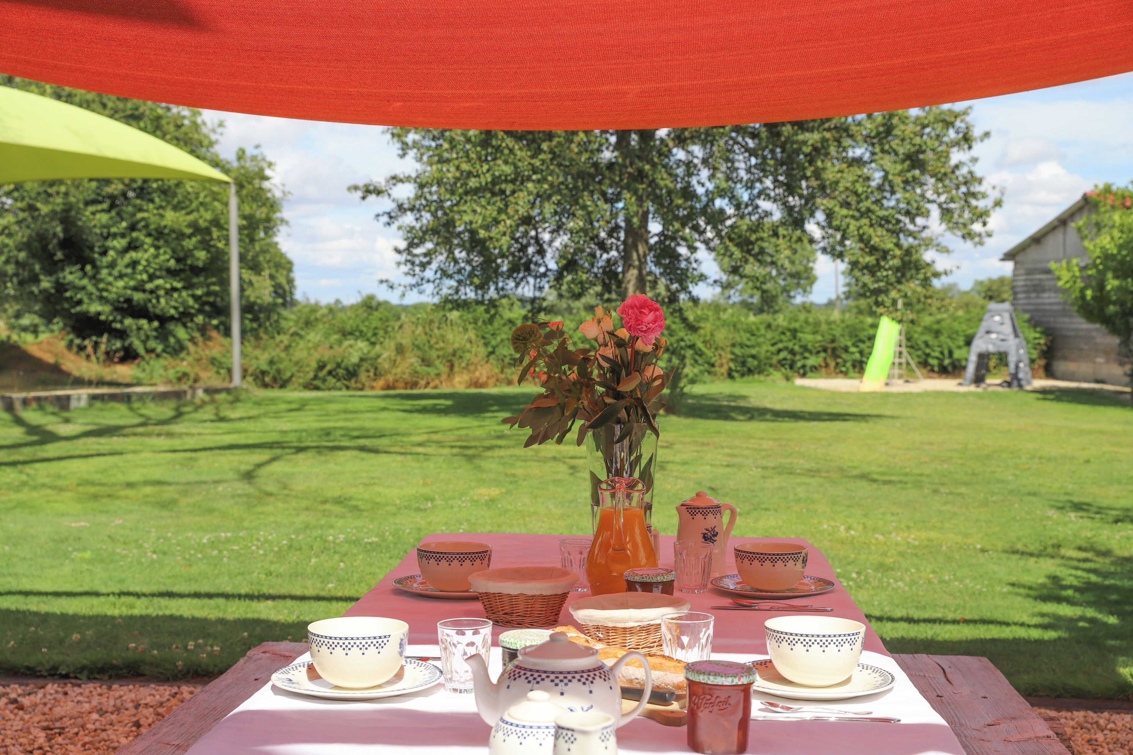 A picnic setup with a table set for tea and snacks, surrounded by greenery and a wooden structure in the background.