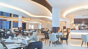 A modern hotel dining area with multiple tables and chairs, a central bar, and a kitchen visible in the background.