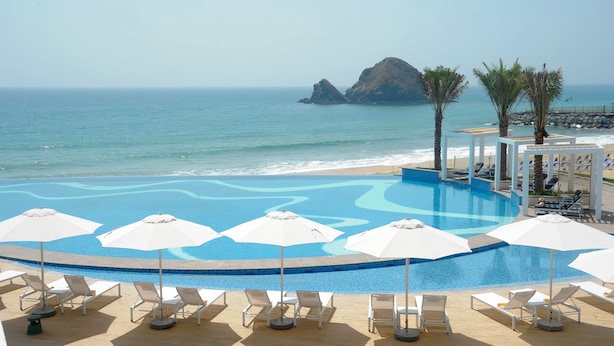 A pool with a wavy design, surrounded by white lounge chairs and umbrellas, with a view of the ocean and a rocky island.