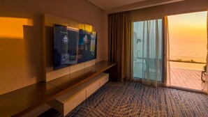 A hotel room with a large TV mounted on the wall, a sliding glass door leading to a balcony with a view of the sea, and a patterned carpet.