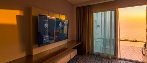 A hotel room with a large TV mounted on the wall, a sliding glass door leading to a balcony with a view of the sea, and a patterned carpet.