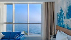 A hotel room with a large window offering a sea view, a bed, a blue sofa, and a painting on the wall.