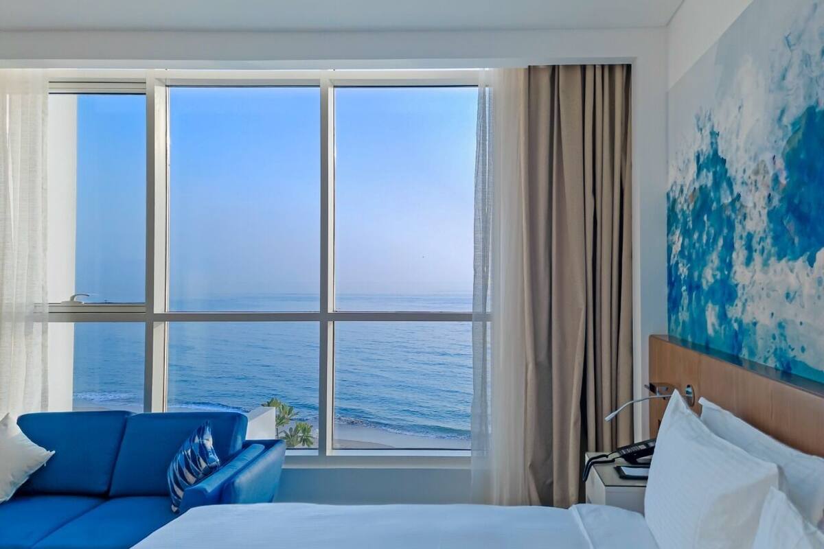 Een hotelkamer met een groot raam met uitzicht op zee, een bed, een blauwe bank en een schilderij aan de muur.
