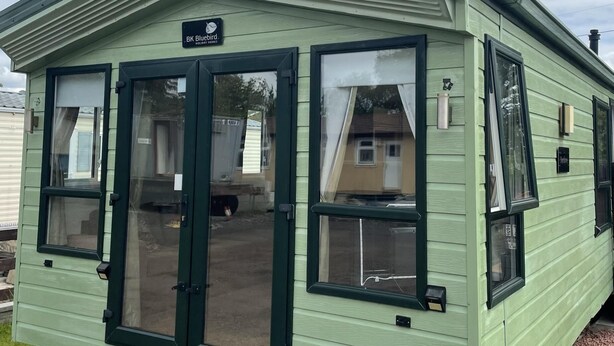 Une petite maison mobile d’un seul étage, de couleur vert clair, avec un toit en pente et une enseigne indiquant « BK Bluebird Holiday Homes ».