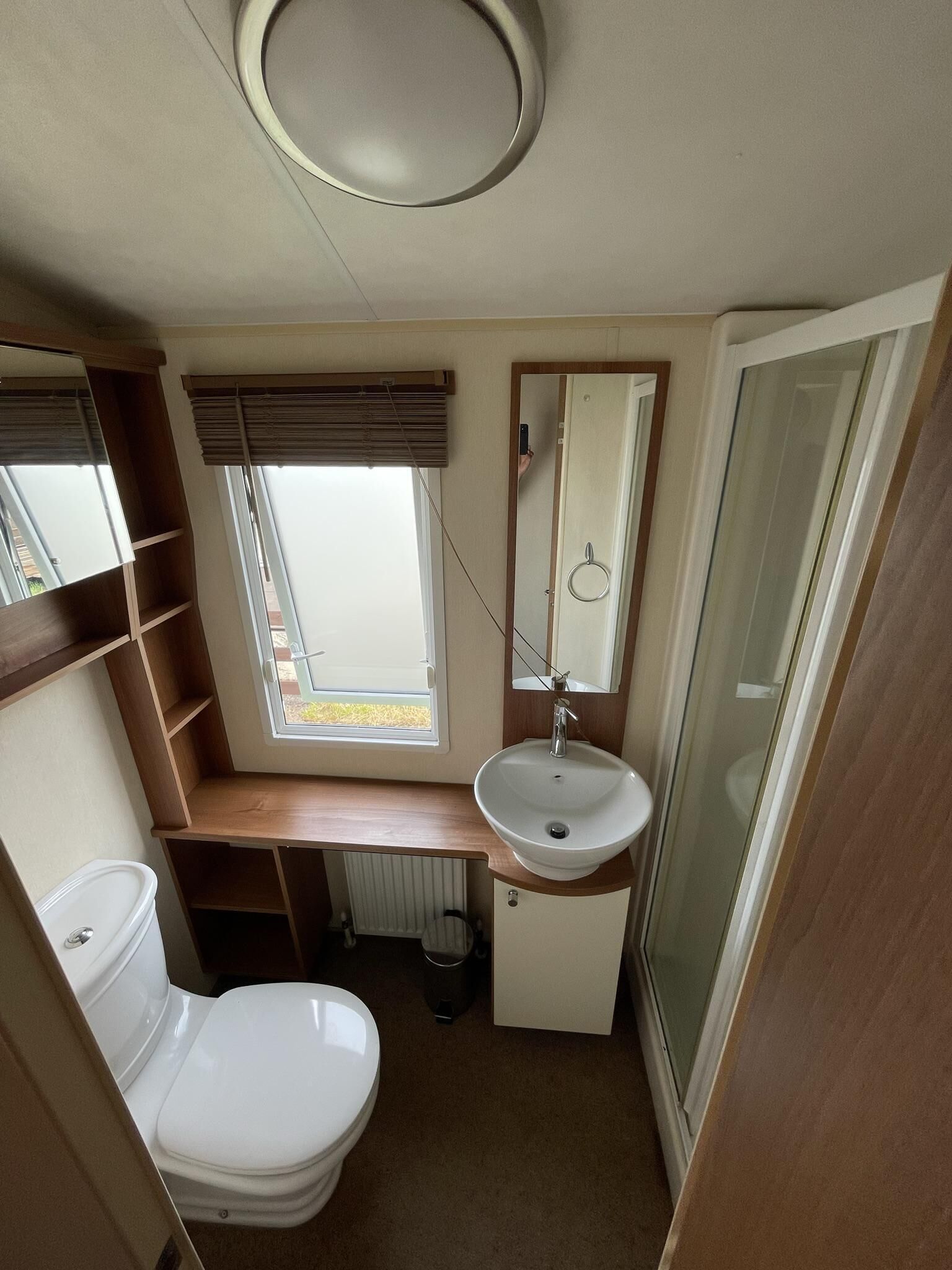 A compact bathroom with a toilet, sink, and mirror. There is a window with blinds, a radiator, and a shower area.