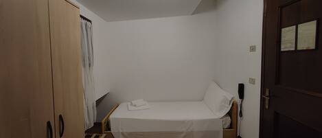A small, sparsely furnished room with a single bed, wooden wardrobe, and a door.