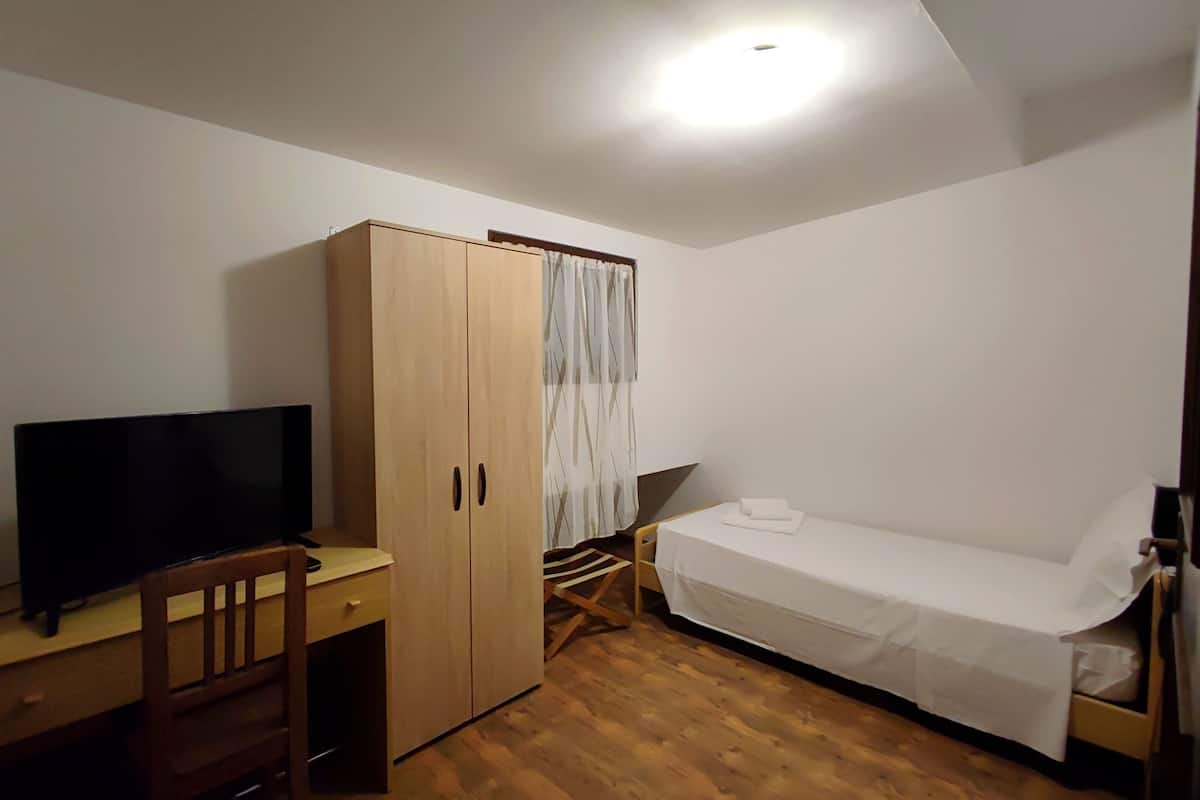 A small, sparsely furnished room with a wooden desk, a television, a wardrobe, and a single bed.