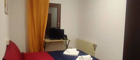 Una stanza piccola con un letto, una scrivania, una televisione e un termosifone.